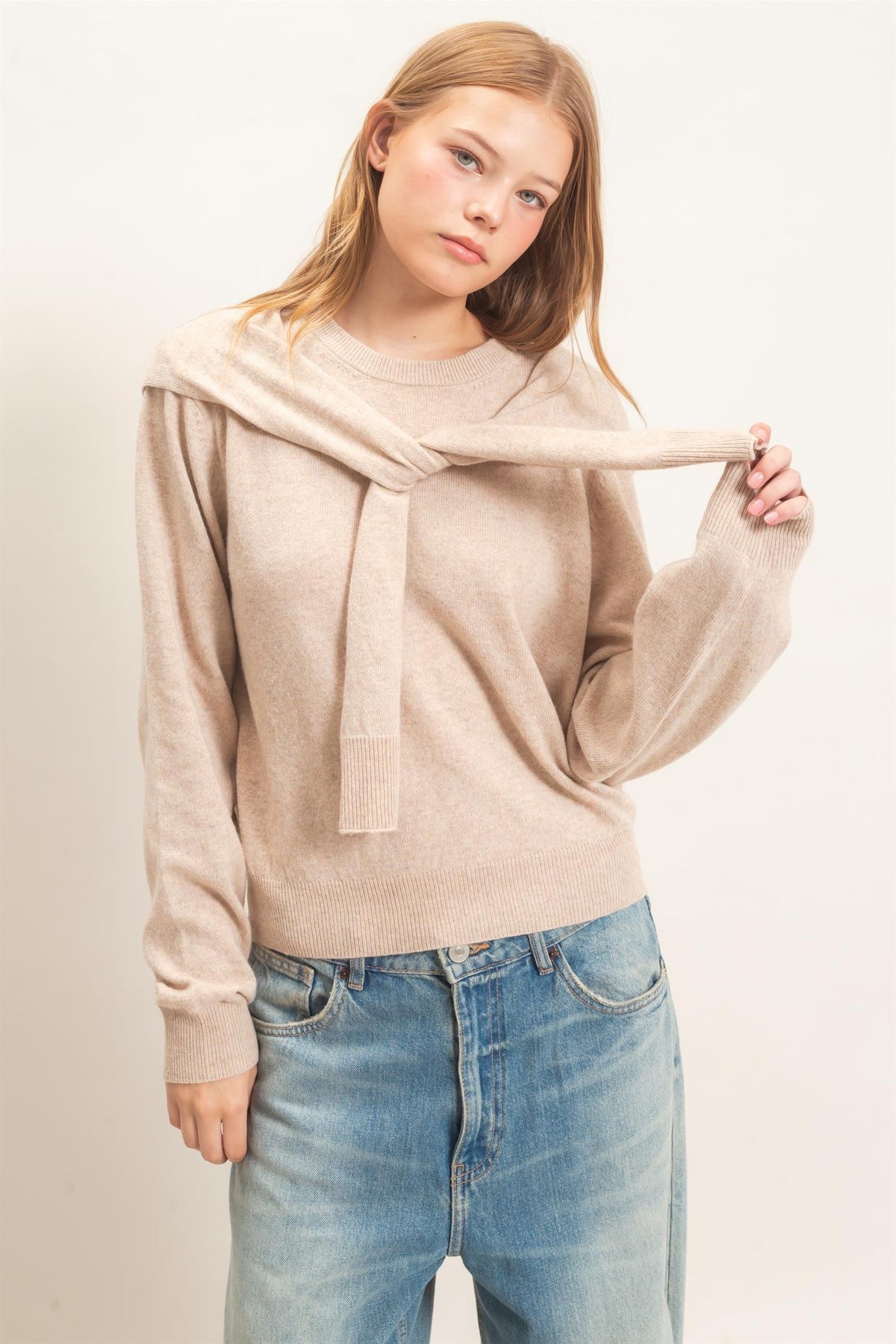 woman standing on white backdrop wearing beige sweater and jeans 