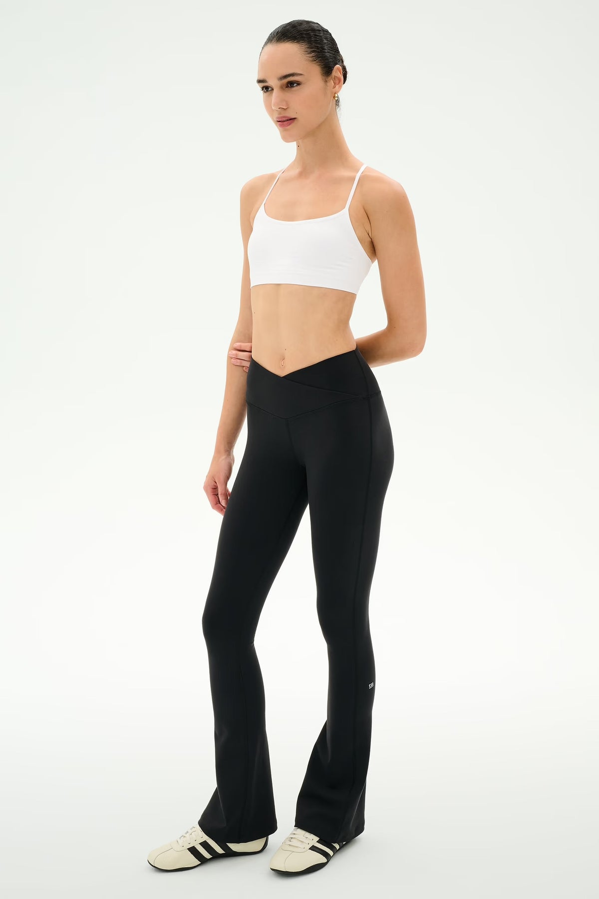 A model wearing black flared leggings, a white sports bra, and white sneakers with black stripes, standing against a plain white studio background.