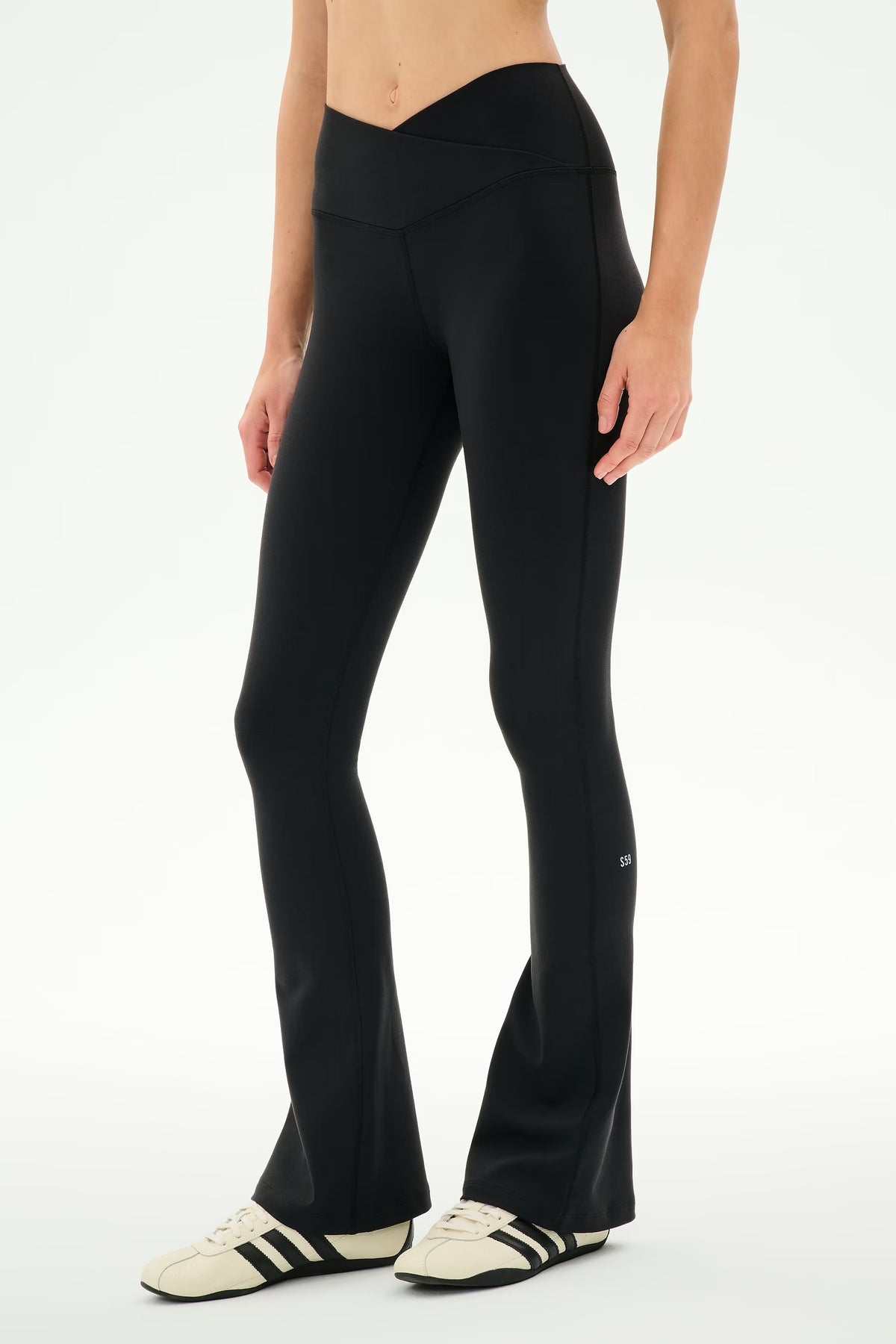 A model wearing black flared leggings and cream sneakers with black stripes, shown from the waist down against a plain white studio background.