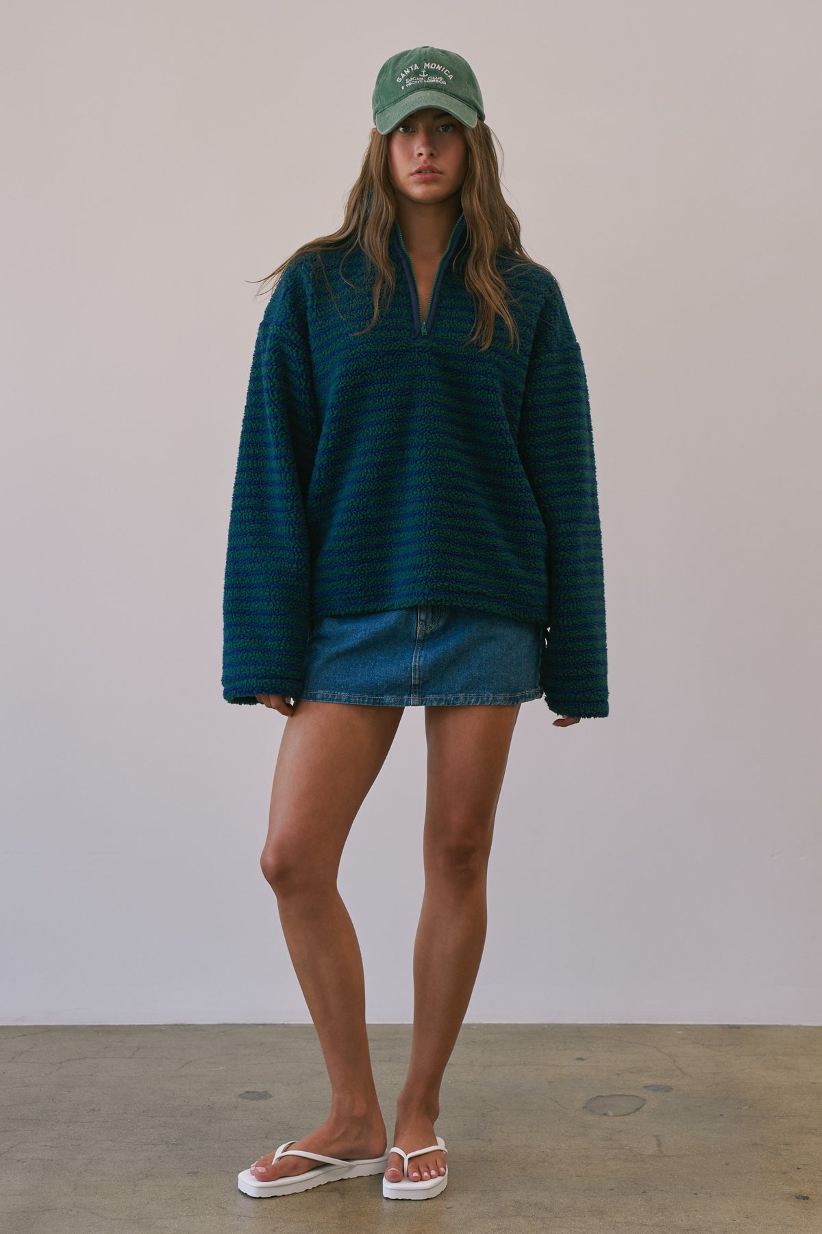 A model stands against a plain light gray wall wearing an oversized dark teal-green ribbed knit half-zip sweater, a denim mini skirt, a green baseball cap, and white flip-flop sandals.