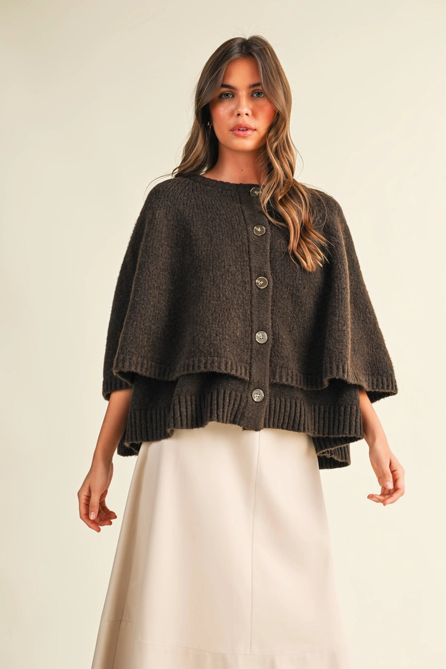 A woman modeling a dark brown textured knit cape cardigan with button-front closure and a cream-colored skirt, set against a plain beige studio background.