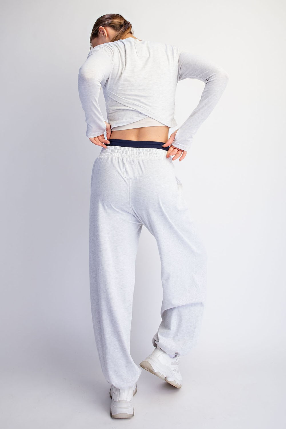 A woman shown from behind wearing a light gray heathered two-piece loungewear set consisting of a long-sleeve crop top and matching sweatpants, paired with white sneakers against a light gray studio background.