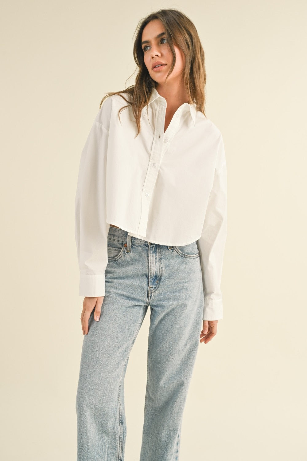 Woman standing on tan background wearing a cropped white button front shirt and jeans