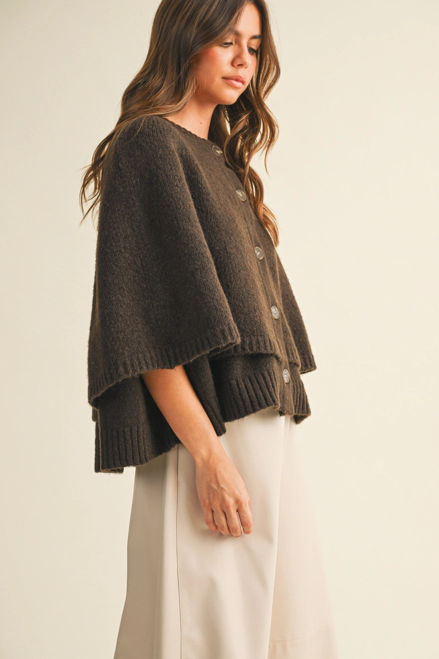 A woman wearing a dark brown knit poncho cardigan with wide draped sleeves and front buttons, paired with light beige wide-leg pants, against a plain cream background.