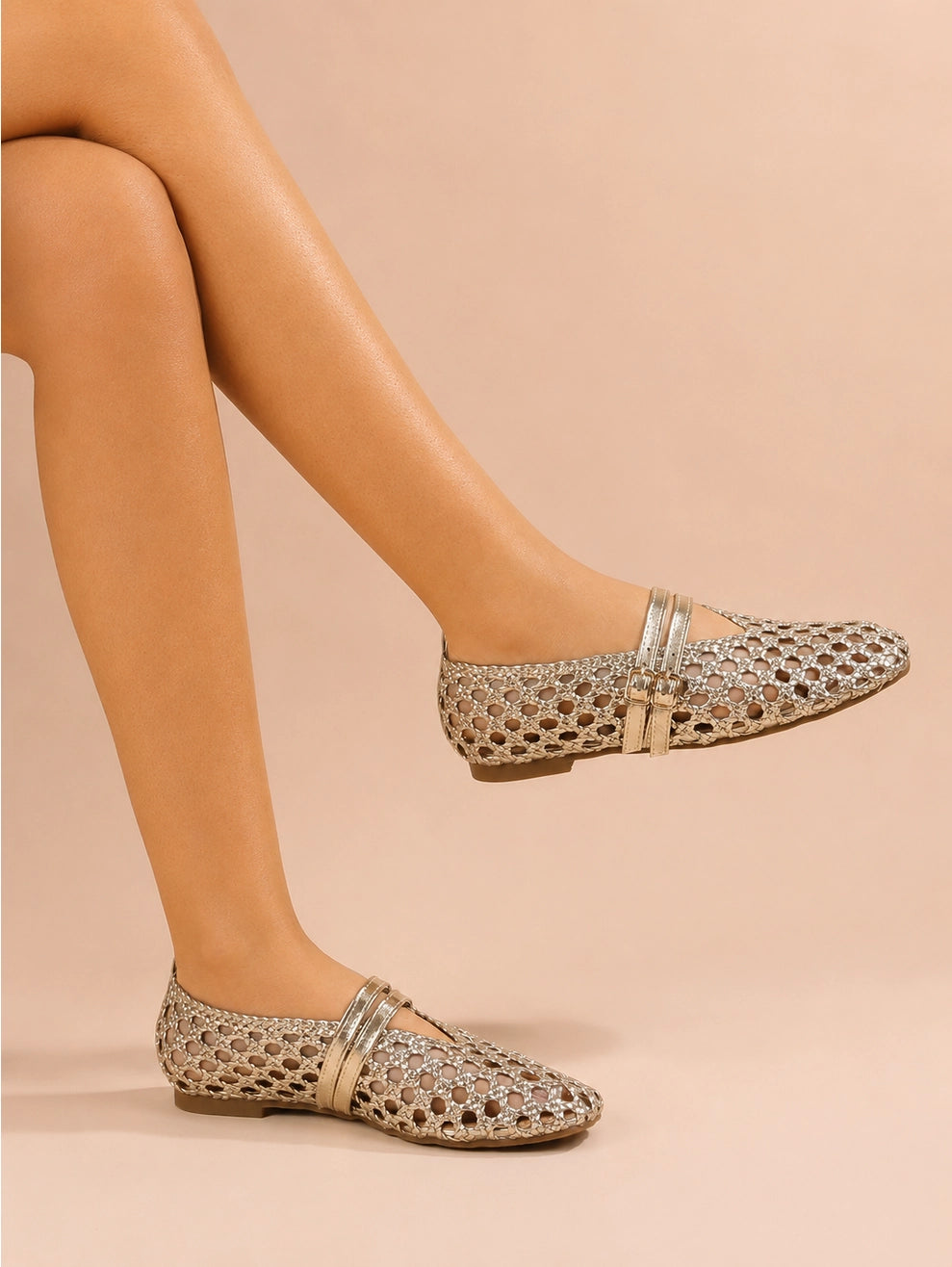 A pair of women's openwork flat shoes with metallic gold cutout detailing and a gold strap with a gold buckle, shown on a model's feet against a solid beige background.