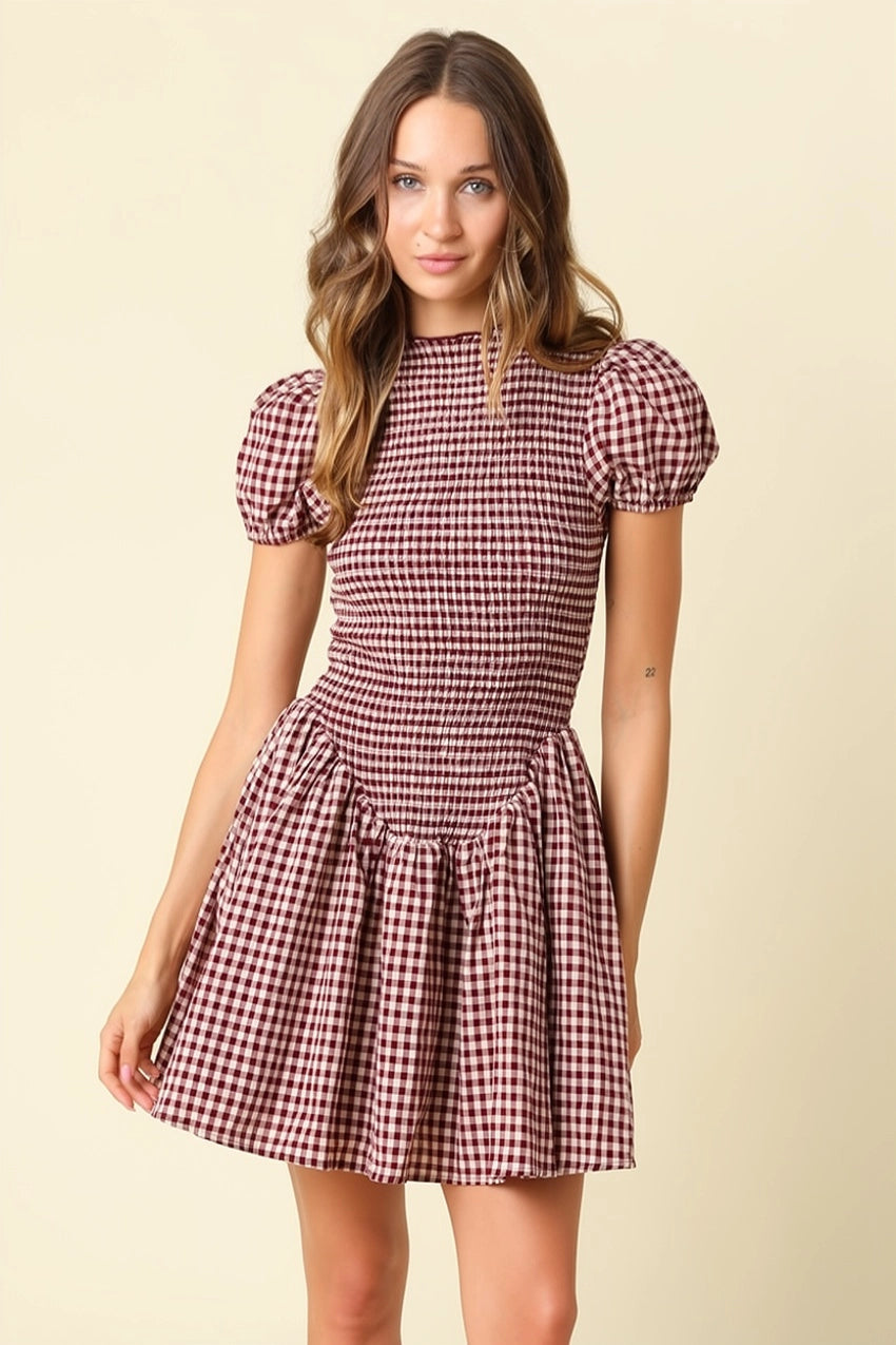 A woman modeling a short puff-sleeve dress with a red and white gingham check pattern against a solid light beige background.