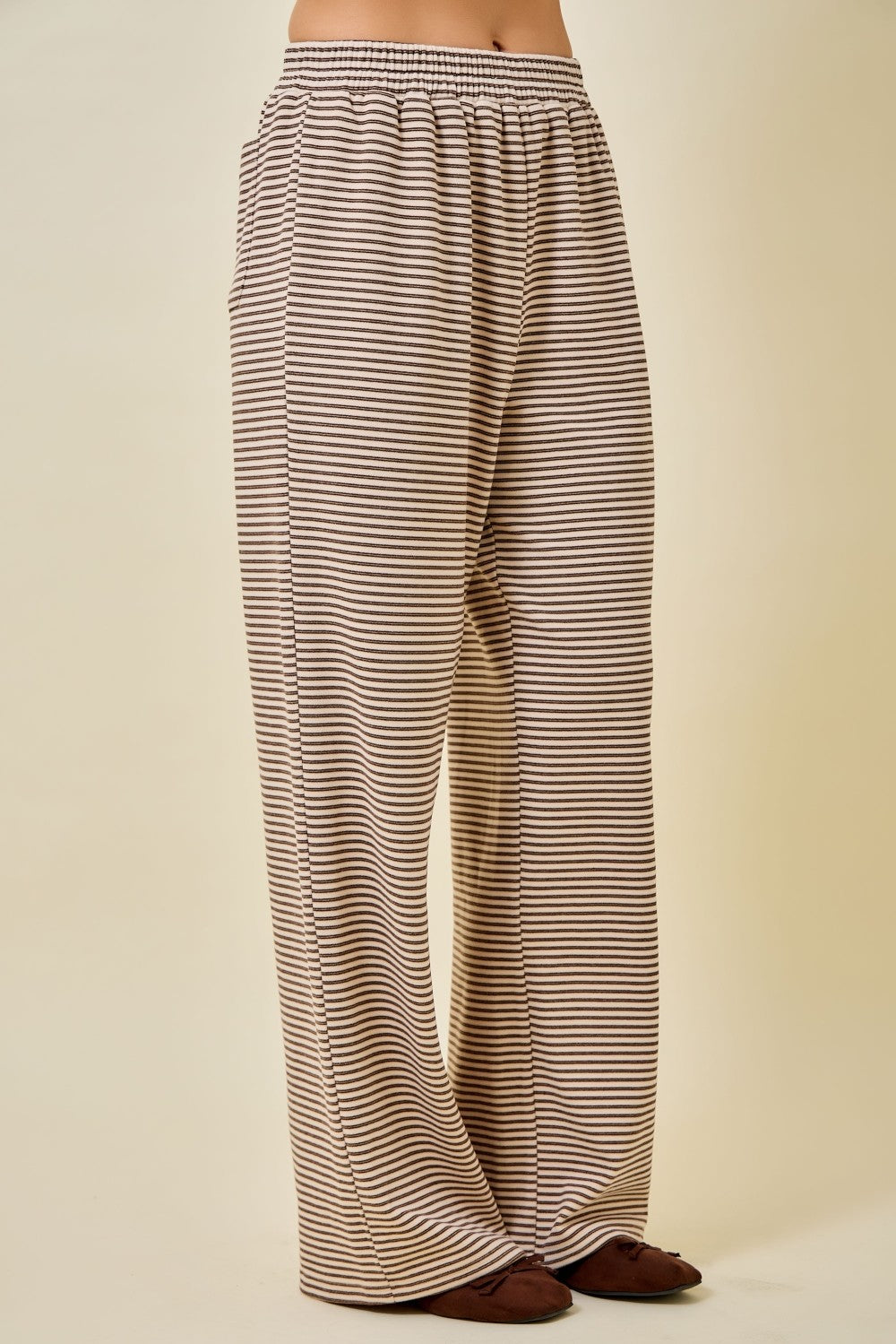 A person wearing loose, wide-leg pants with thin horizontal brown and beige stripes, shown from the waist down against a plain beige studio background.