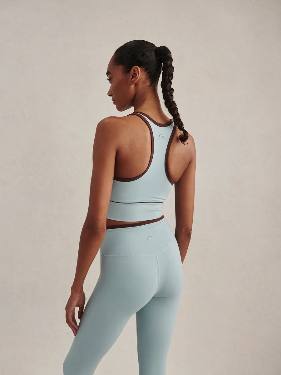 A woman shown from the back wearing a light blue athletic set consisting of a sports bra and leggings, with dark brown trim, against a neutral beige studio background.
