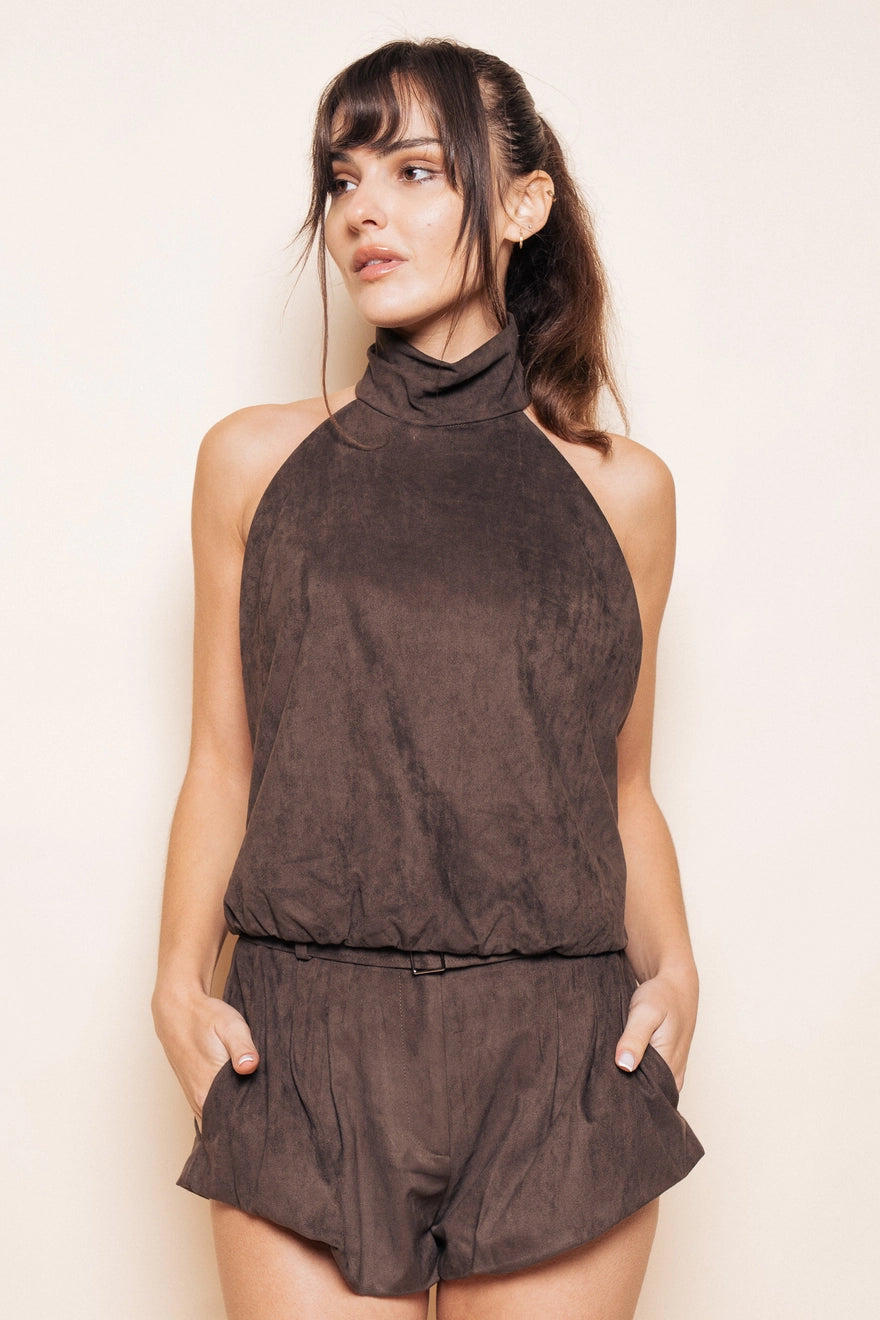 Model with brown hair wearing a brown suede halter tank top with matching brown bubble shorts with pockets standing in front of a beige background. 