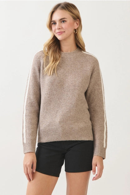 A woman wearing a light taupe knit sweater with white stripes down the sleeves, paired with black shorts, against a plain white studio background.