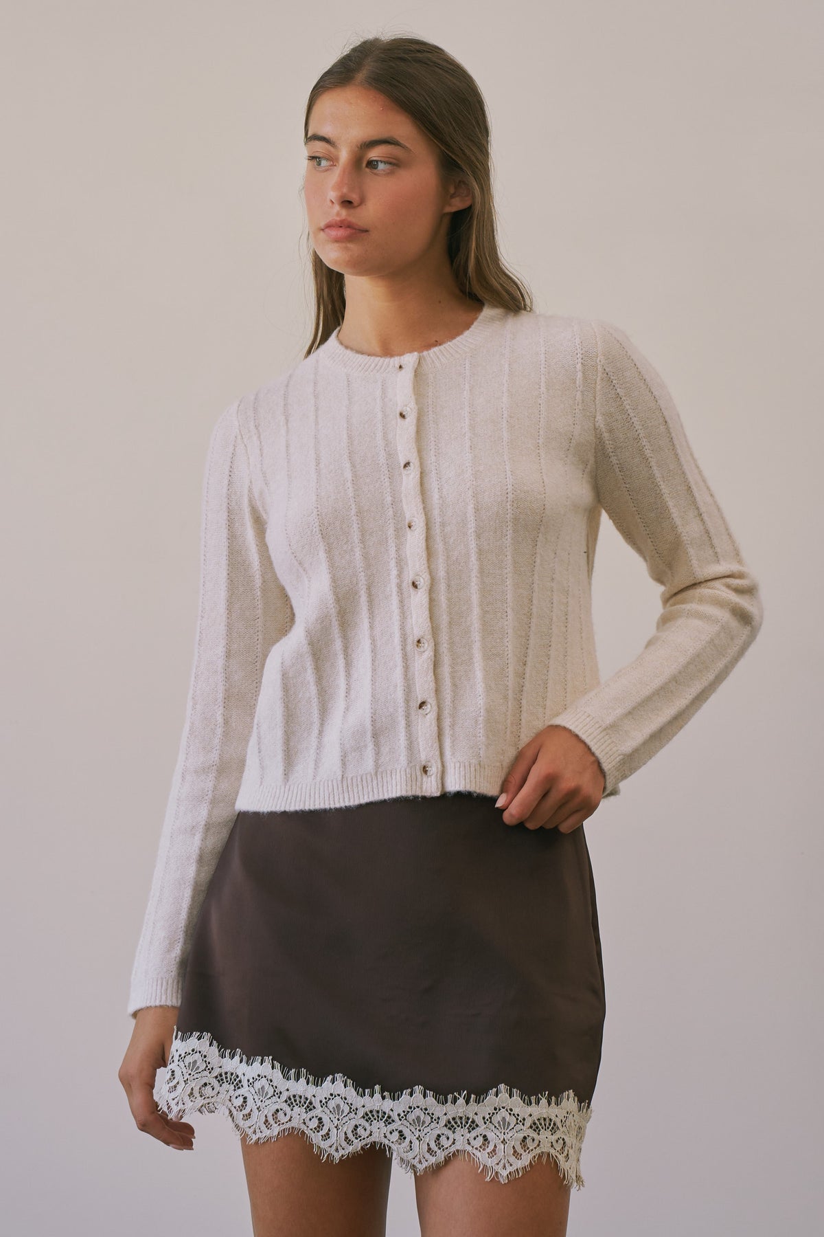 A woman modeling a cream ribbed knit cardigan with buttons over a dark brown skirt with white lace trim, set against a plain light beige studio background.