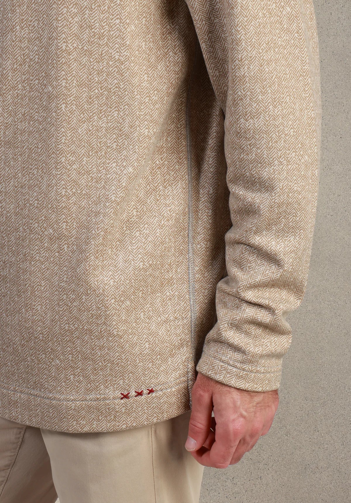 Close-up of a person wearing a beige textured knit sweater with a fine herringbone weave, paired with matching beige trousers. The sweater features a ribbed hem and cuffs, with a small red embroidered detail near the bottom hem.