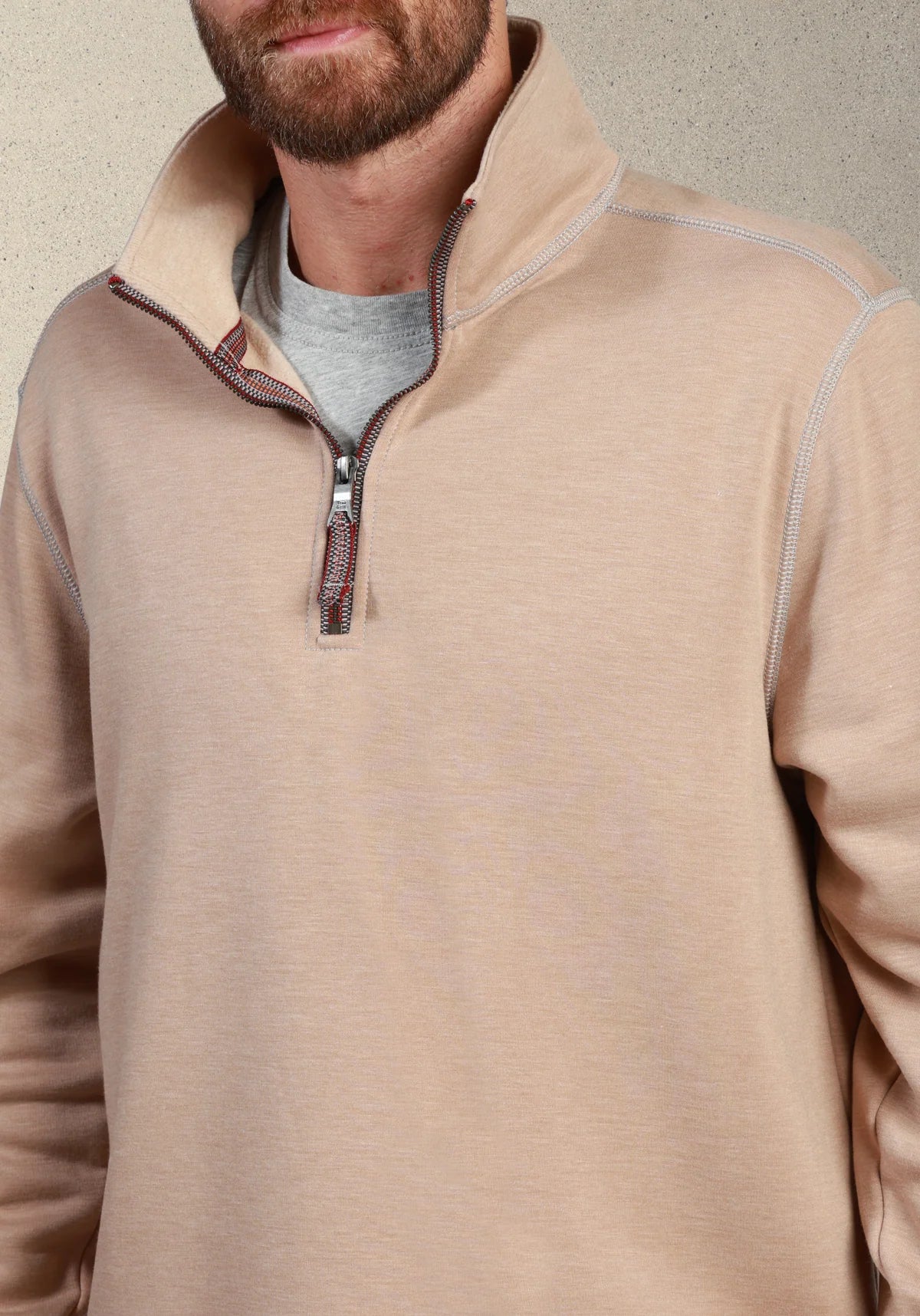 A close-up of a man wearing a beige quarter-zip pullover sweater over a gray t-shirt, set against a neutral textured background.