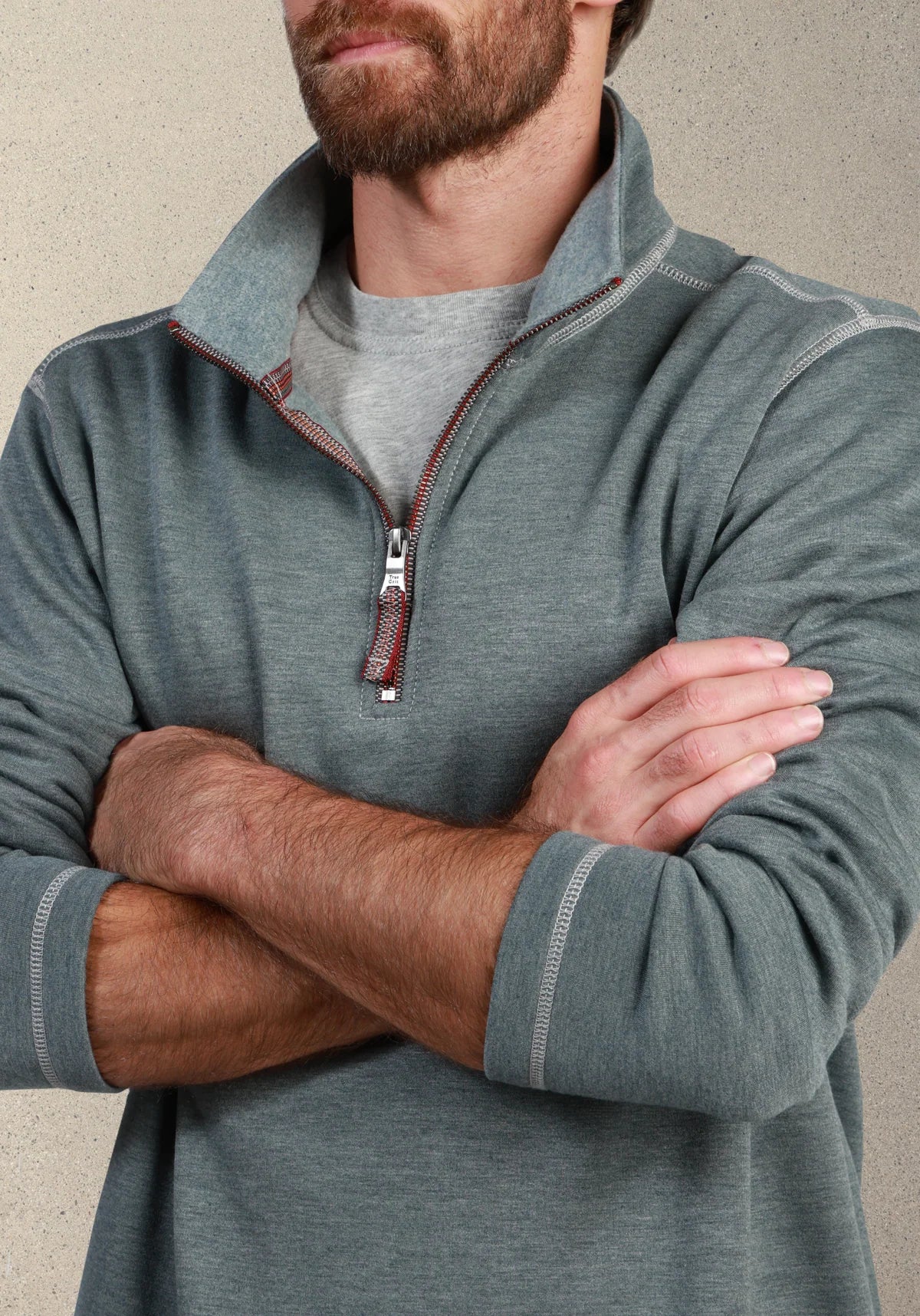 A man wearing a heathered slate gray quarter-zip pullover sweater over a light gray t-shirt, with his arms crossed against a neutral textured background.