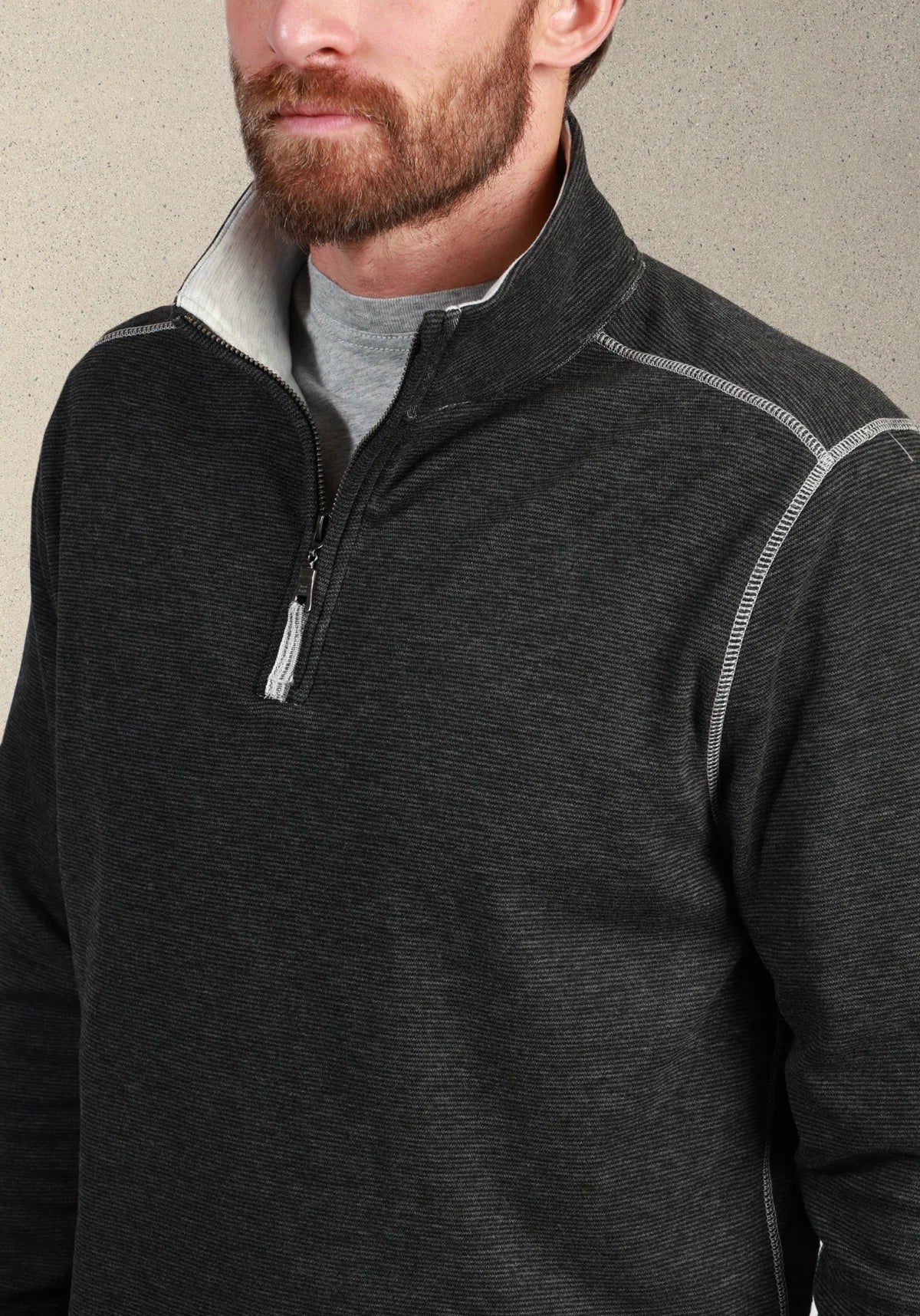 A close-up of a man wearing a dark charcoal gray quarter-zip pullover sweater with a light gray inner collar and a gray t-shirt underneath, set against a neutral textured background.