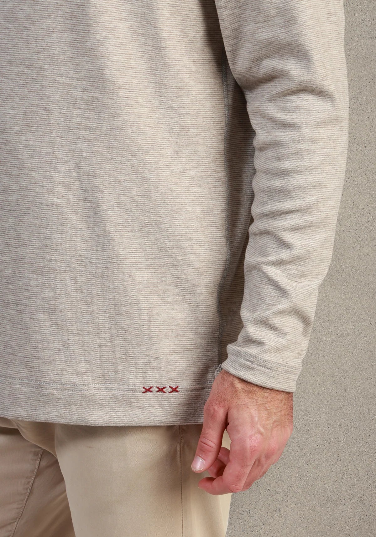 Close-up of a person wearing a light beige long-sleeve knit top with a small red embroidered logo near the hem, paired with beige pants against a neutral studio background.