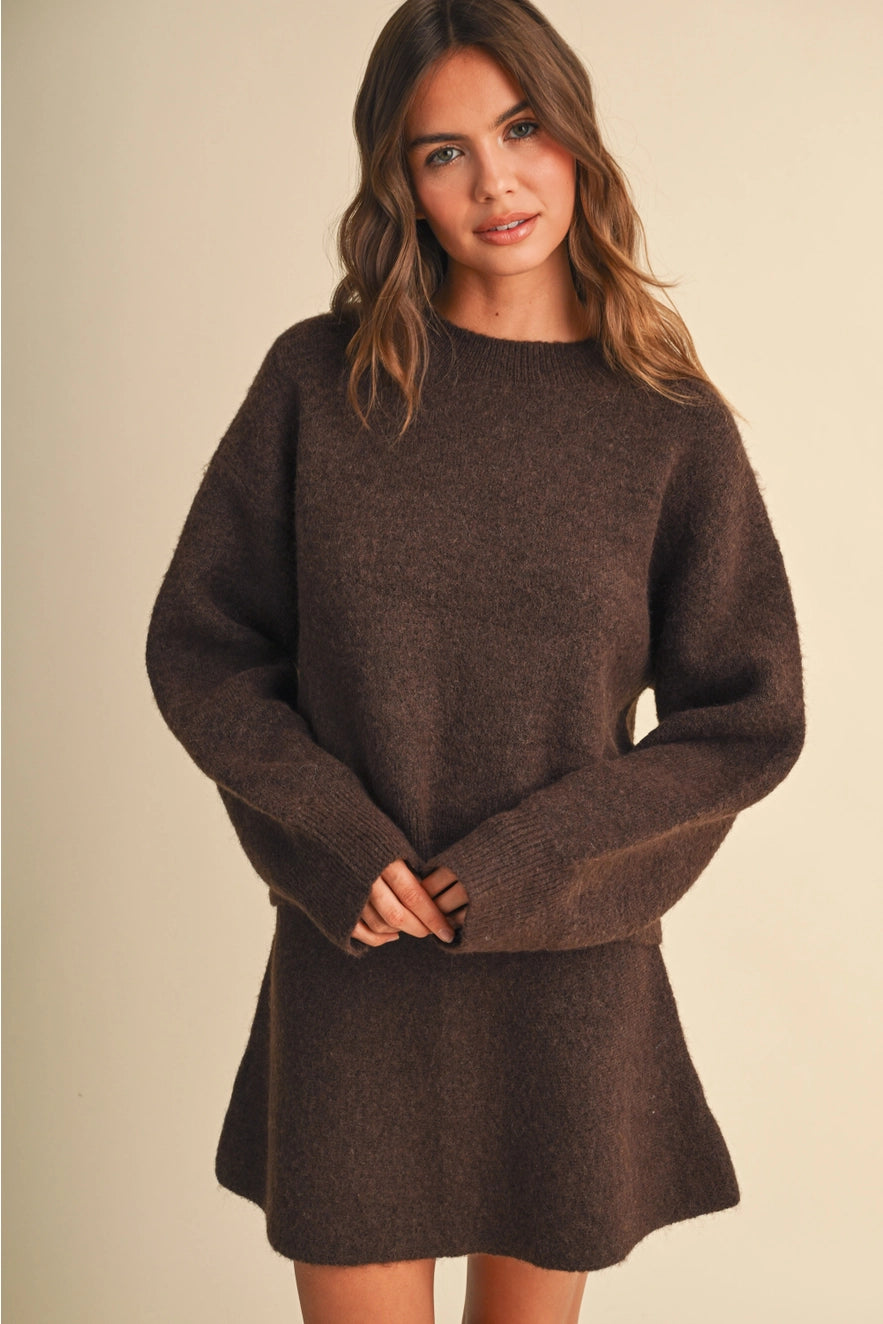 A woman wearing a dark brown oversized knit sweater dress, standing against a solid beige studio background.