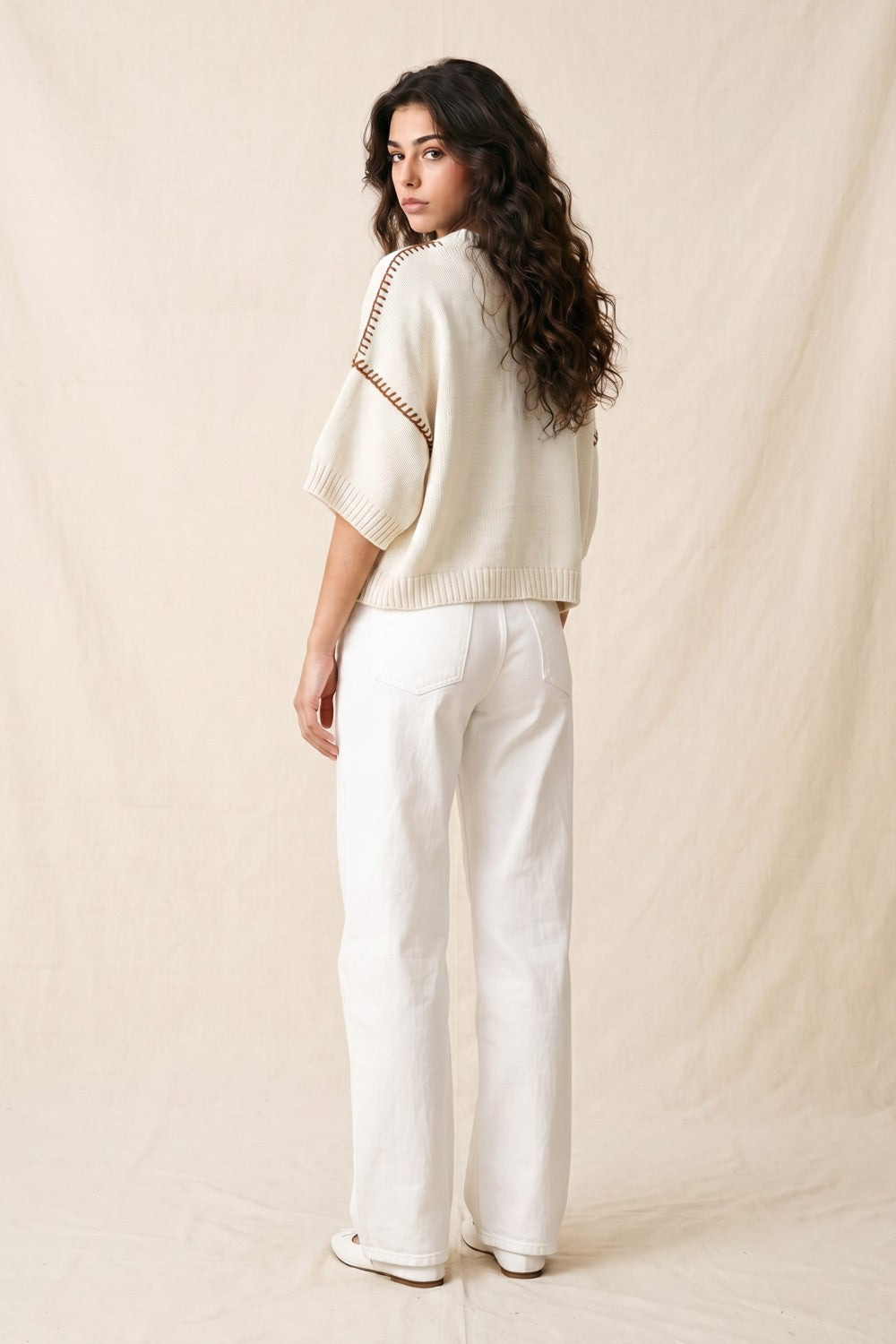 Woman wearing a cream sweater and white pants against a beige background