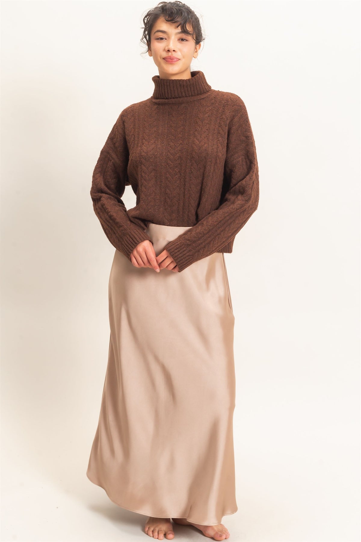 Person wearing a brown turtleneck sweater and beige skirt on a plain background