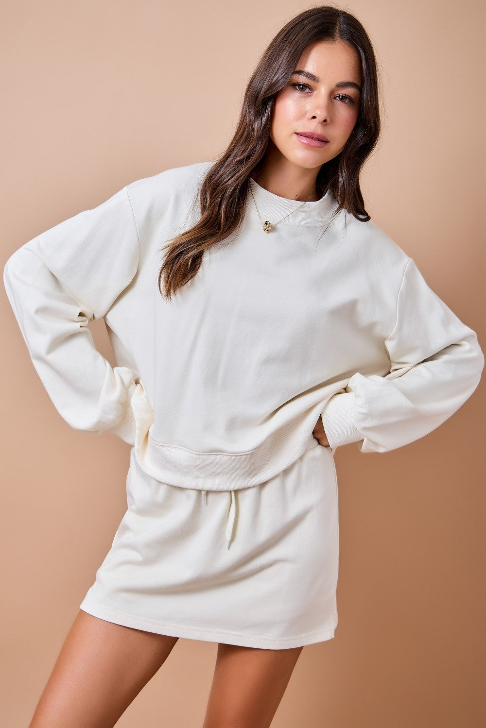 A woman modeling a cream-colored sweatshirt and matching sweat shorts set against a solid beige studio background.