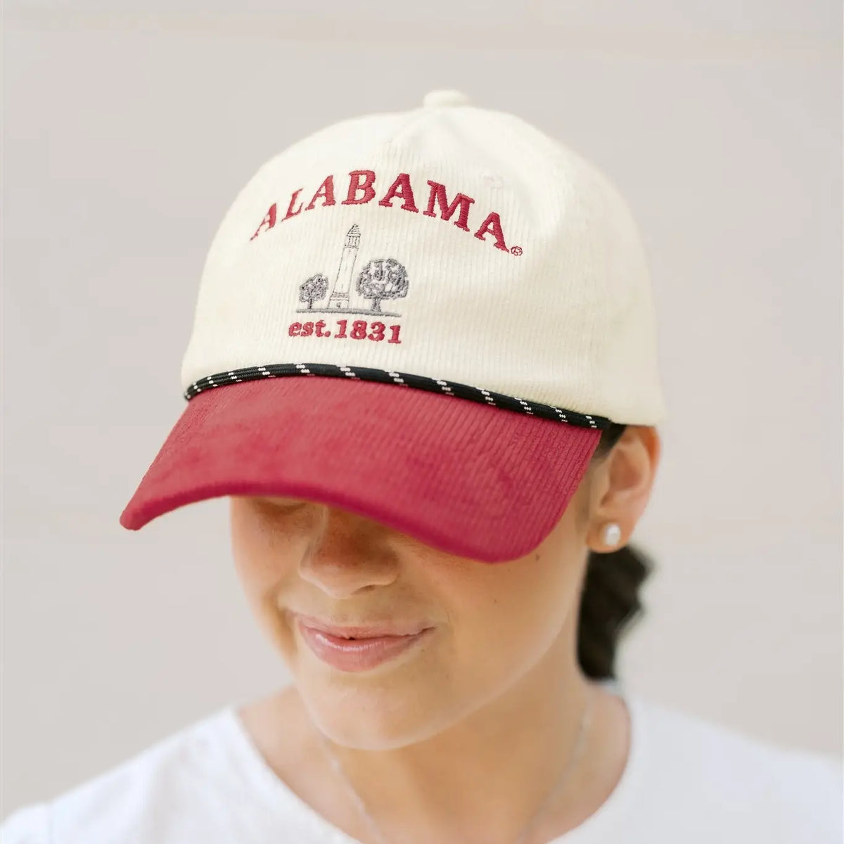 A person wearing a cream-colored baseball cap with red embroidered 'ALABAMA' text and a maroon visor, photographed against a light neutral background.