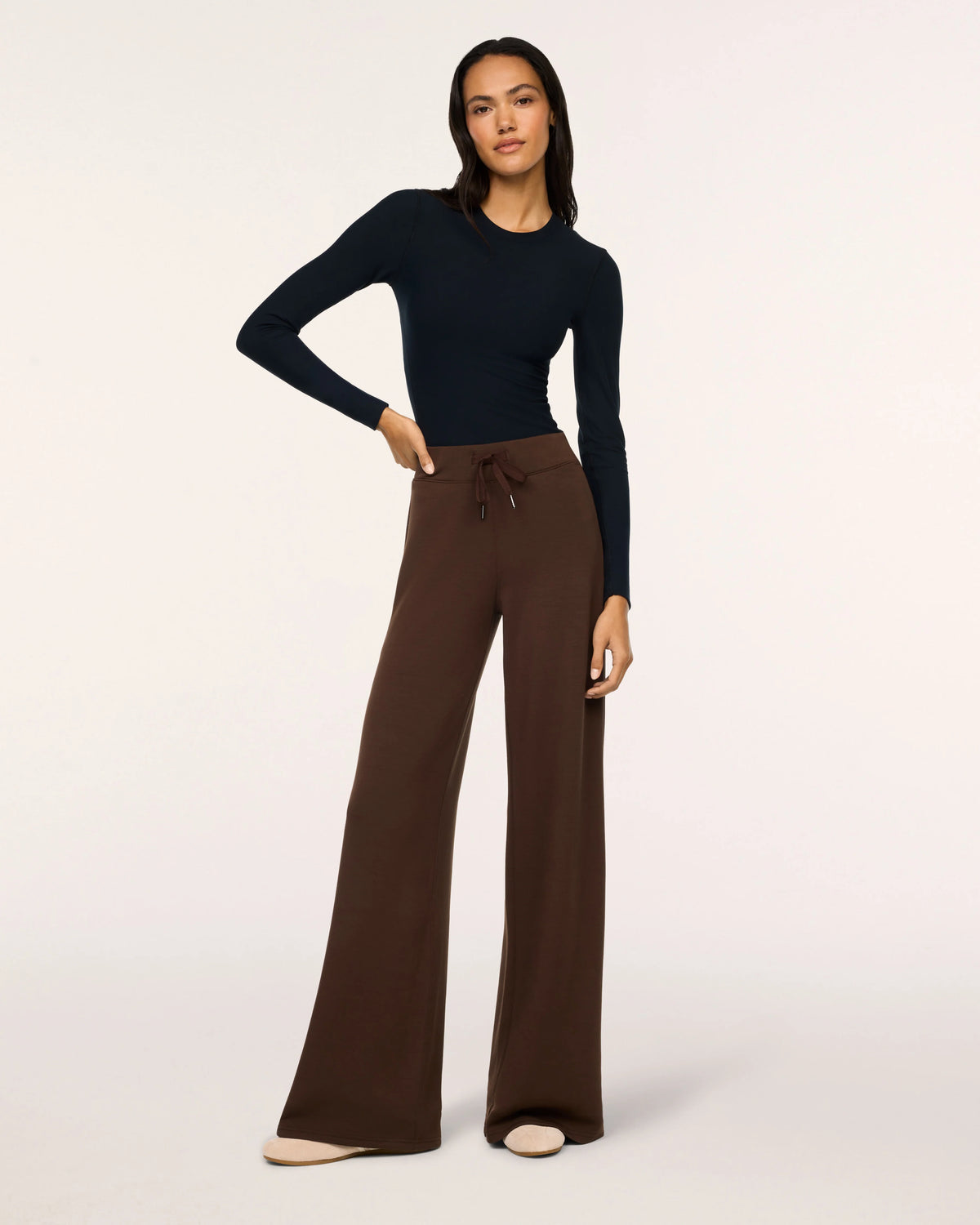 A model wearing a black long-sleeve mock-neck top and brown wide-leg drawstring pants, standing against a plain off-white studio background.