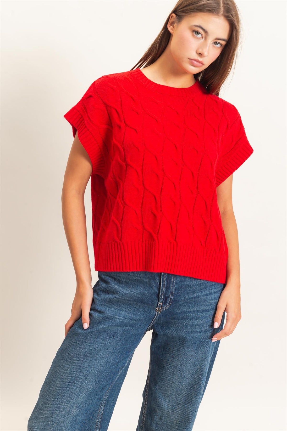 A woman wearing a bright red short-sleeve cable-knit sweater and blue jeans, photographed against a plain light background.