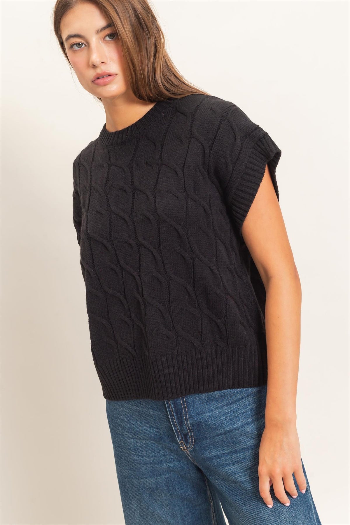 A woman wearing a black cable-knit short-sleeve sweater and blue jeans, photographed against a plain light beige studio background.