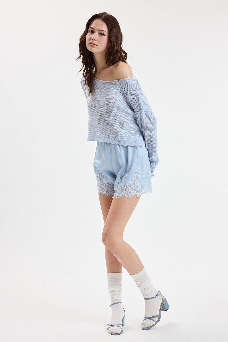 A woman modeling a light blue knit off-the-shoulder sweater and matching light blue lace-trimmed shorts, paired with white socks and light blue heels, against a plain white studio background.