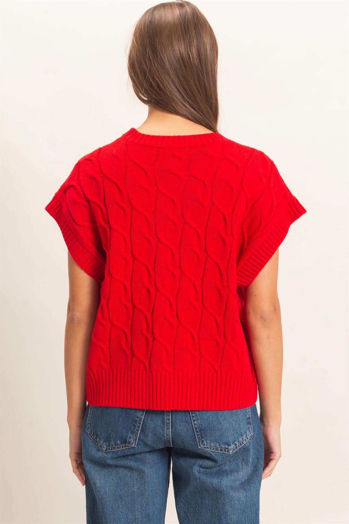 A woman viewed from the back wearing a bright red short-sleeve cable-knit sweater and blue jeans against a plain light background.