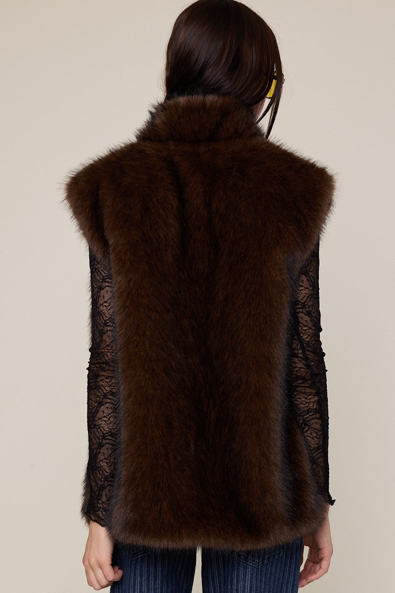 A model shown from the back wearing a dark brown fur vest over a black lace long-sleeve top and blue jeans, against a plain beige background.