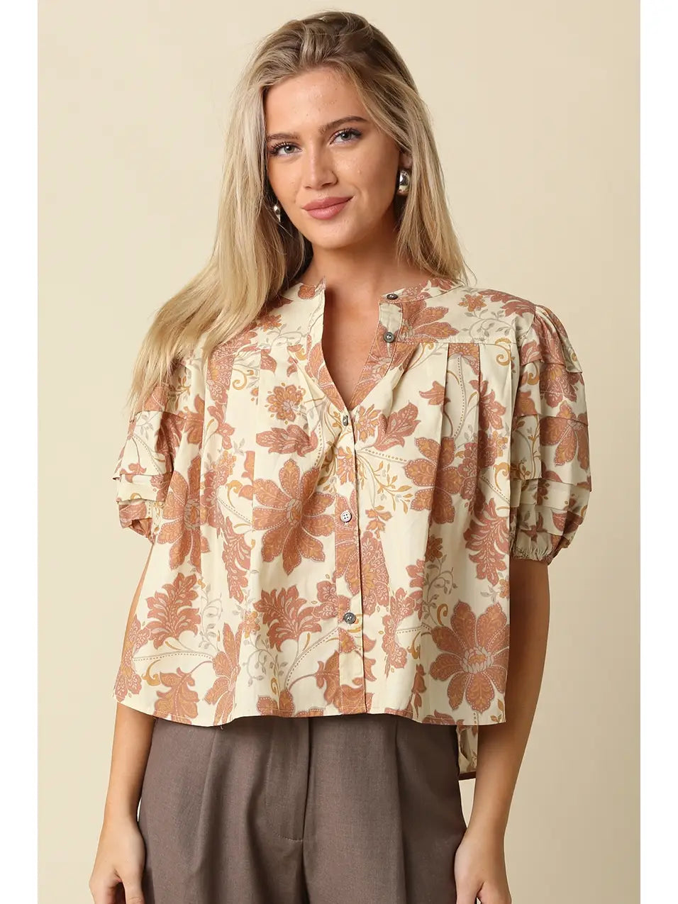 A woman wearing a short-sleeve button-front blouse with a beige and rust floral print, paired with taupe pants, against a solid beige background.