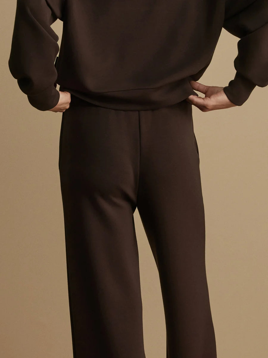 A person wearing a dark brown matching lounge set, featuring a cropped view of a sweatshirt and wide-leg trousers, set against a solid warm beige background.