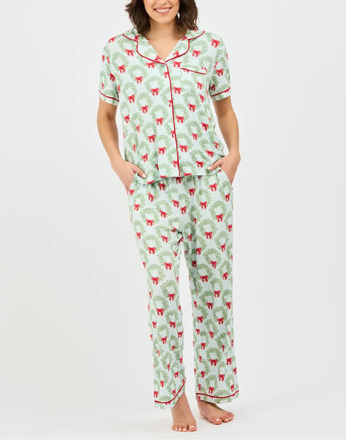 A woman modeling a two-piece pajama set with a light green background, red and green holiday pattern, and red piping on a light gray studio background.