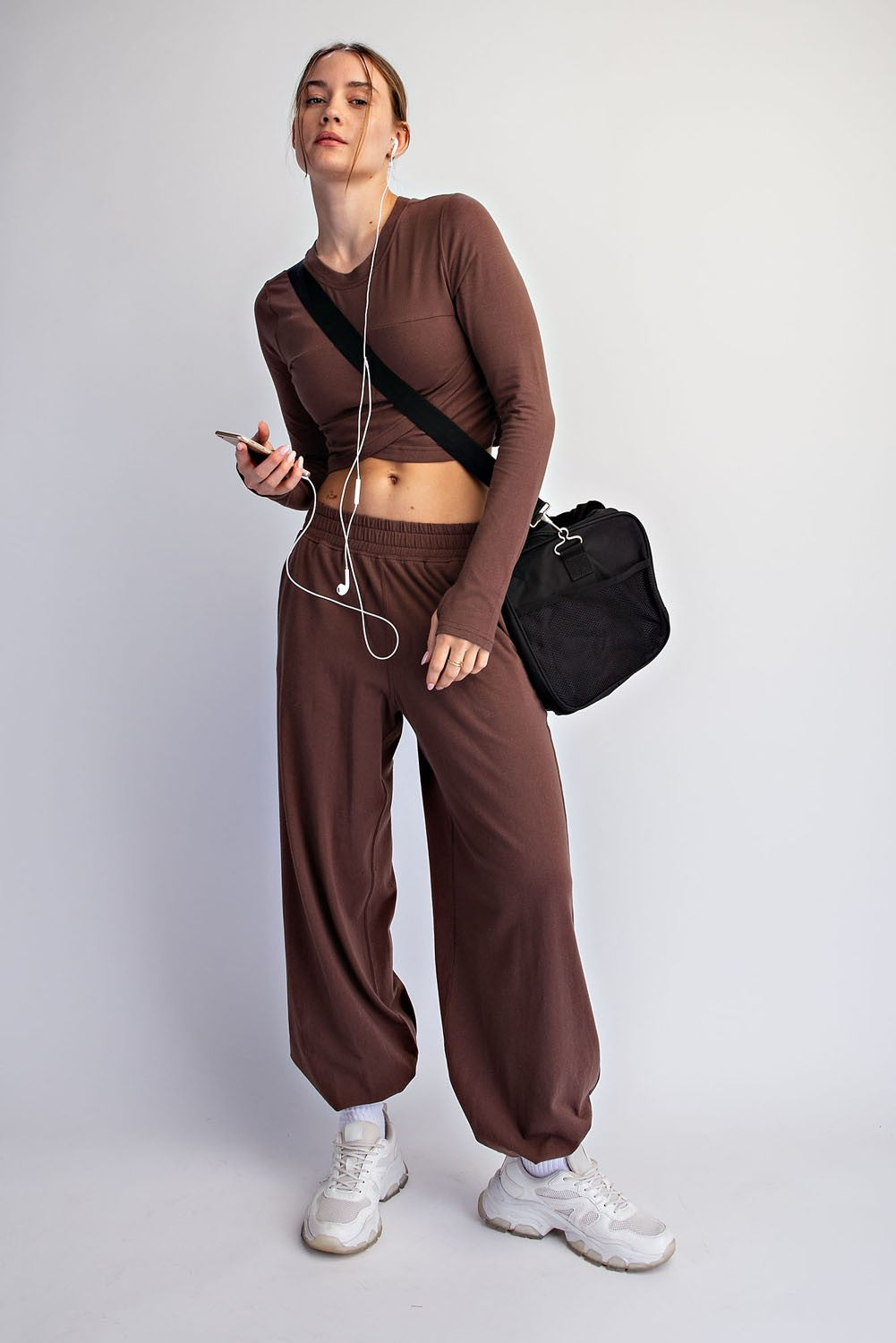 A woman stands against a light gray studio background wearing a matching brown cropped long-sleeve top and jogger-style pants set, with white sneakers, white earphones, and a black shoulder bag.