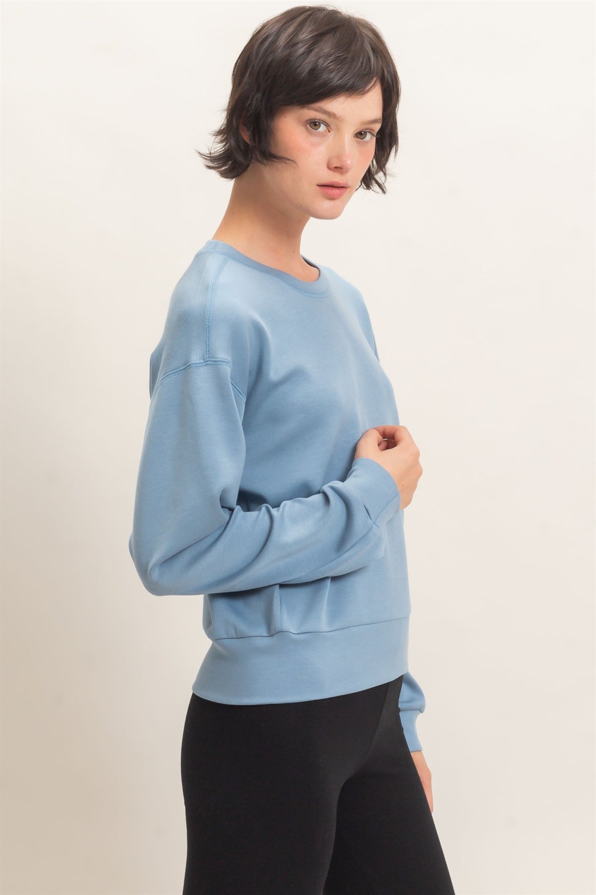 A woman modeling a light blue crewneck sweatshirt with black leggings, photographed against a plain off-white studio background.