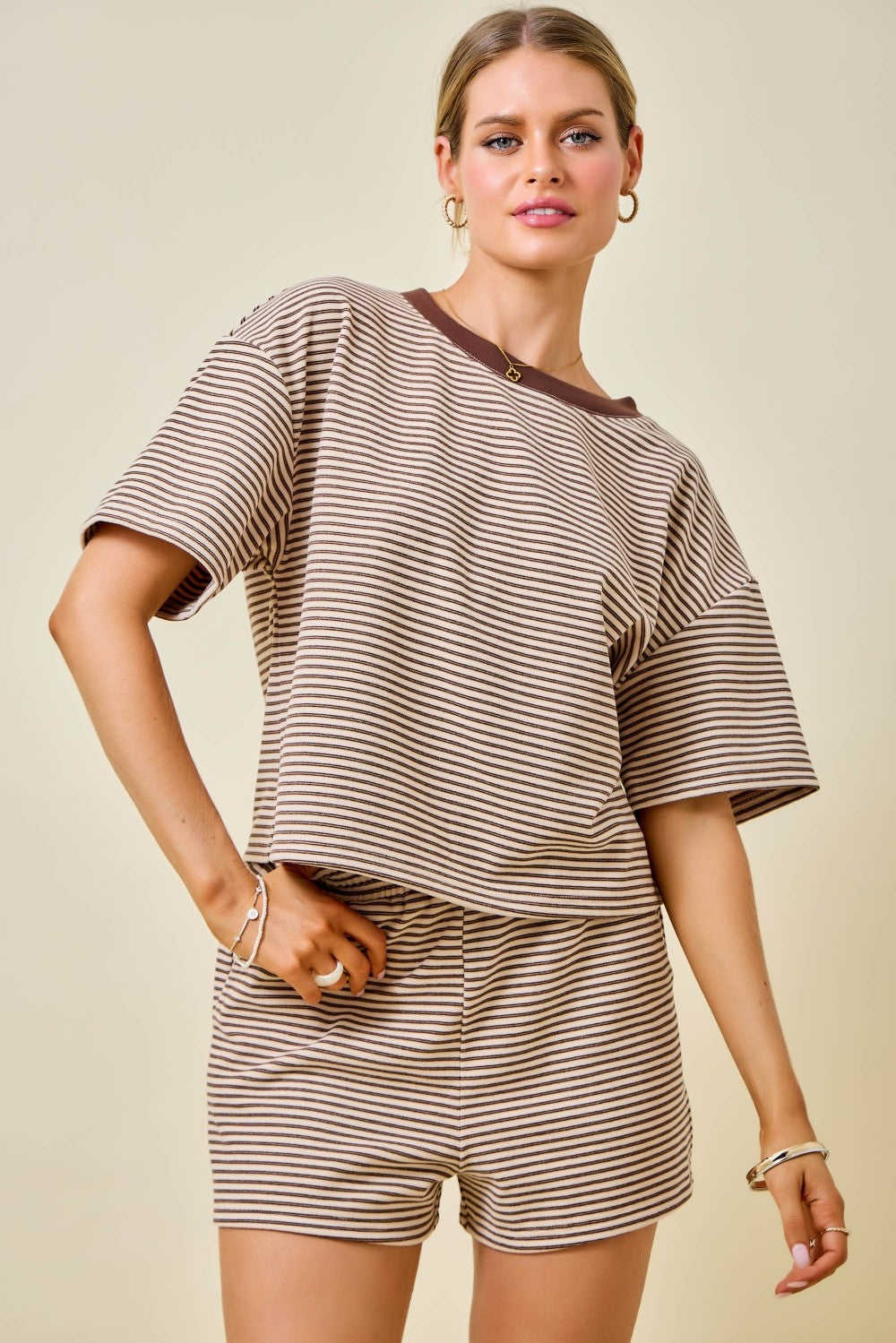 A woman modeling a matching two-piece lounge set featuring a brown and beige striped short-sleeve top with a solid brown collar and matching striped shorts, set against a solid beige background.