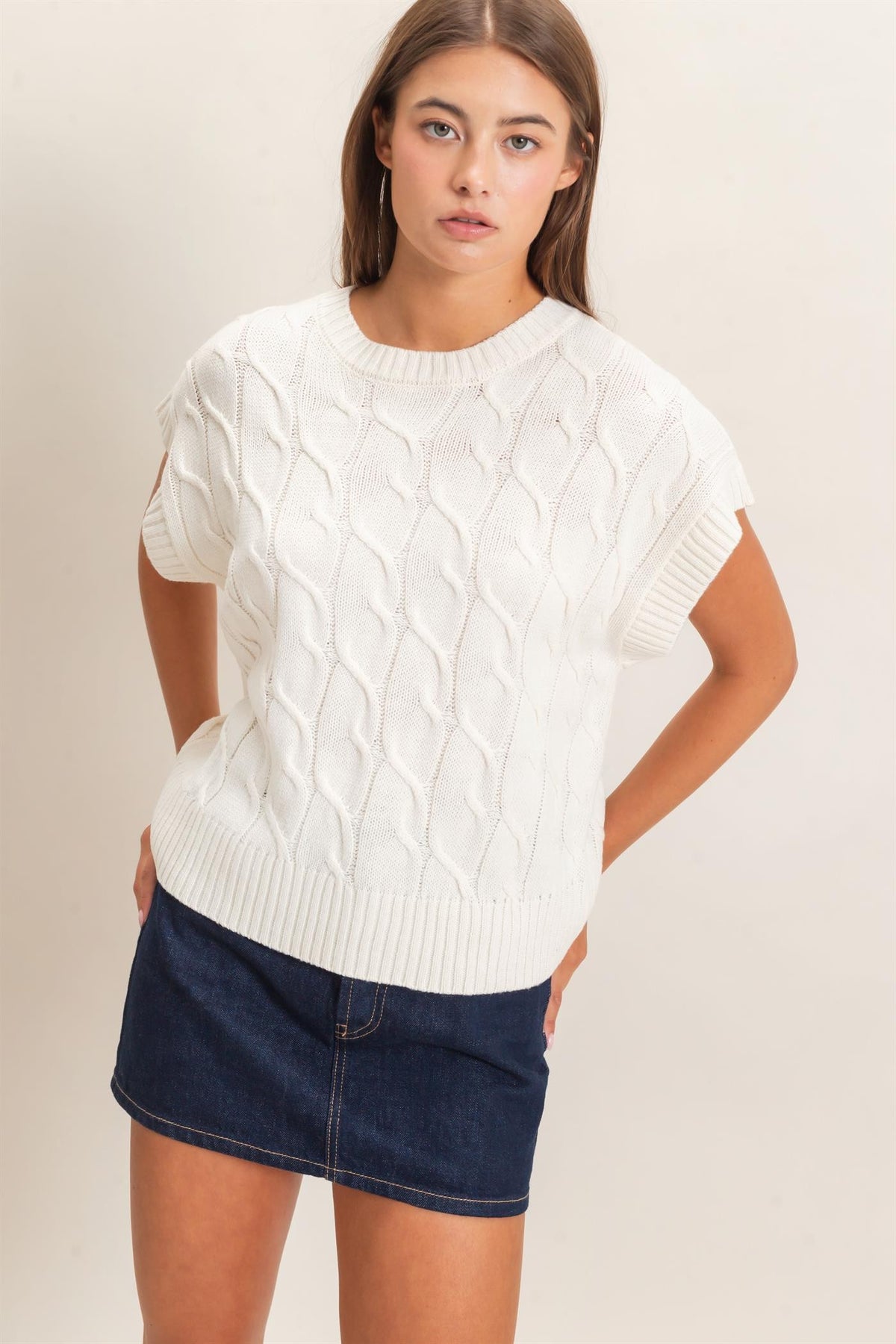 A woman modeling a short-sleeve cable-knit sweater in off-white, paired with a dark denim mini skirt, against a plain beige studio background.