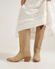 Beige cowboy boots worn with a white dress on a plain background