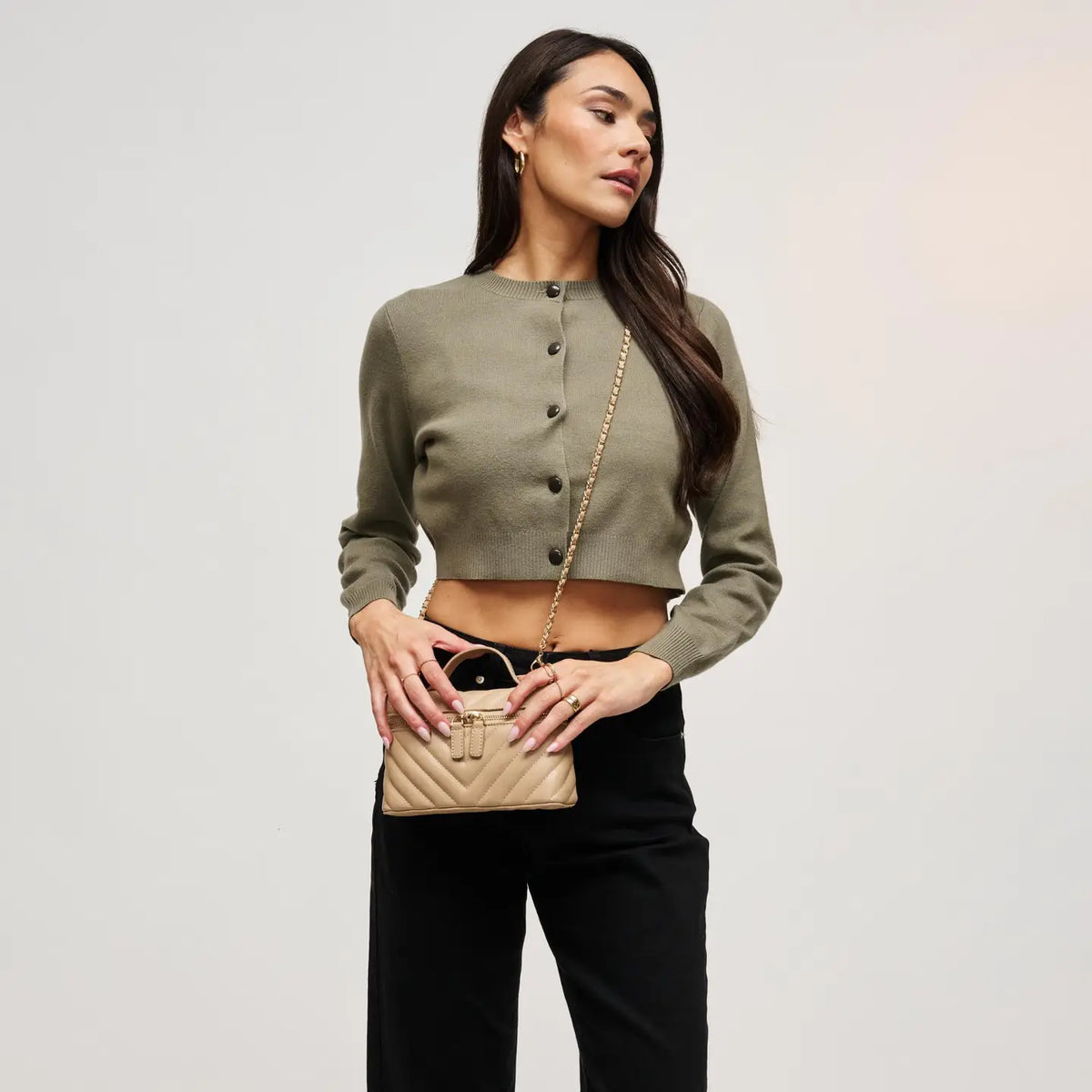 A woman modeling a cropped olive green knit top with front buttons, paired with black pants and holding a small beige quilted handbag with a gold chain strap against a plain light gray studio background.