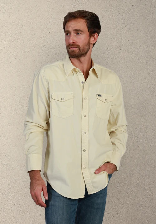 A man wearing a light cream-colored long-sleeve western-style button-down shirt with two chest pockets, paired with blue jeans, standing against a textured beige wall.