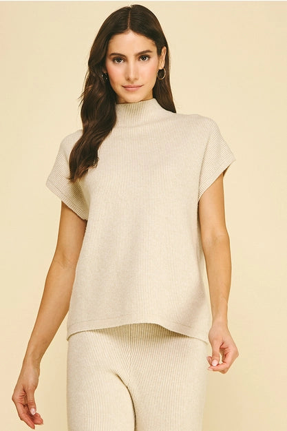A woman modeling a light beige ribbed knit mock neck top with short sleeves, paired with matching ribbed knit pants, against a solid light beige background.