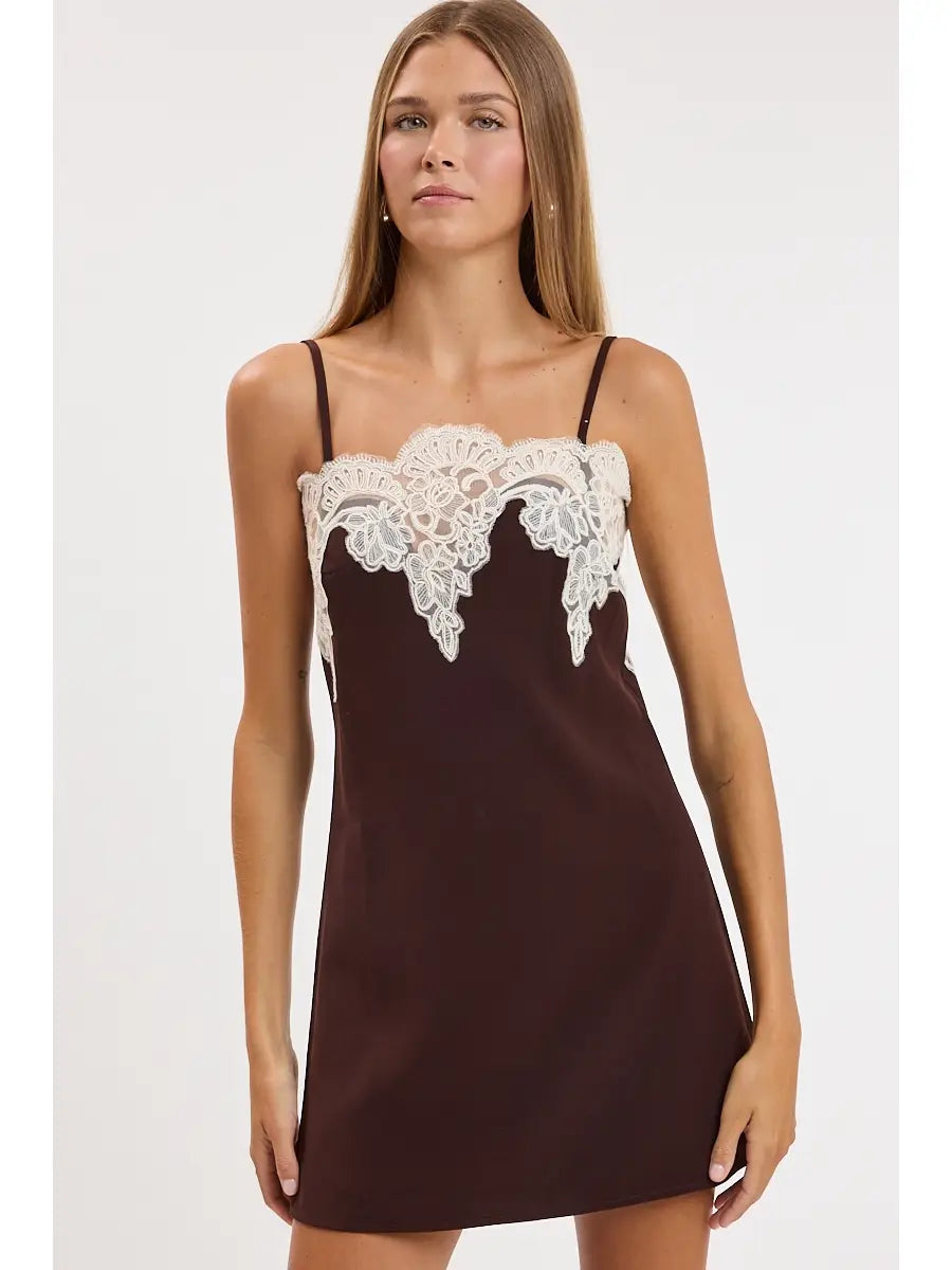 A woman modeling a dark brown satin slip dress with white lace trim across the bust, photographed against a plain light gray studio background.