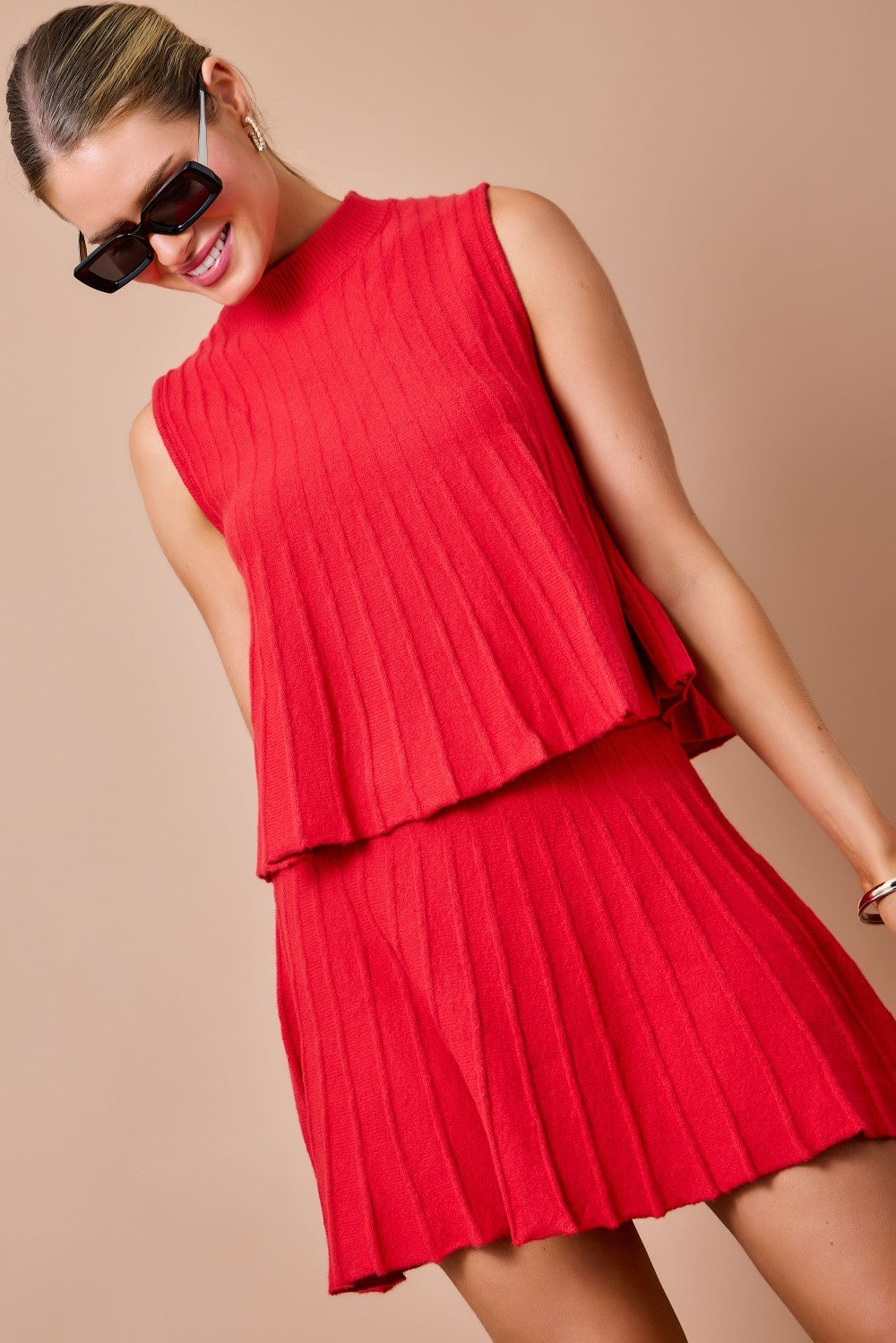A woman wearing a bright red sleeveless pleated mini dress with a layered peplum-style top and matching pleated skirt, posed against a plain beige studio background.