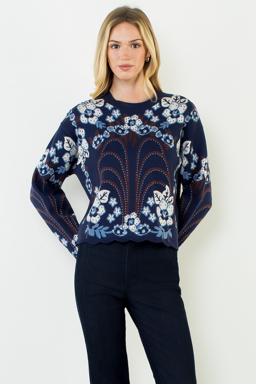 blonde woman standing in dark jeans modeling a navy sweater with floral pattern across the whole thing including sleeves. with a scallop hem