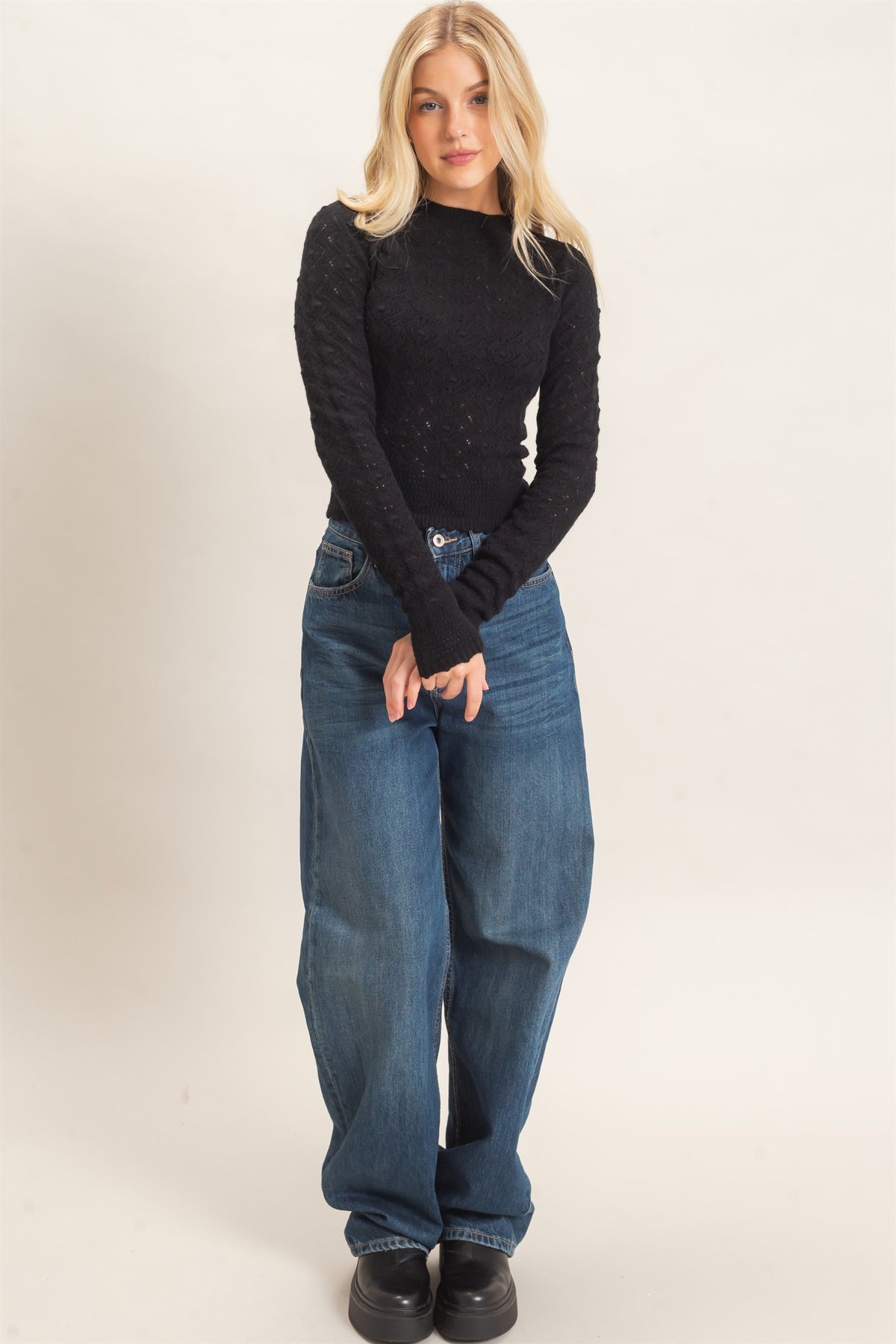 A blonde woman modeling a black long-sleeve knit top and wide-leg blue jeans against a plain light beige studio background.