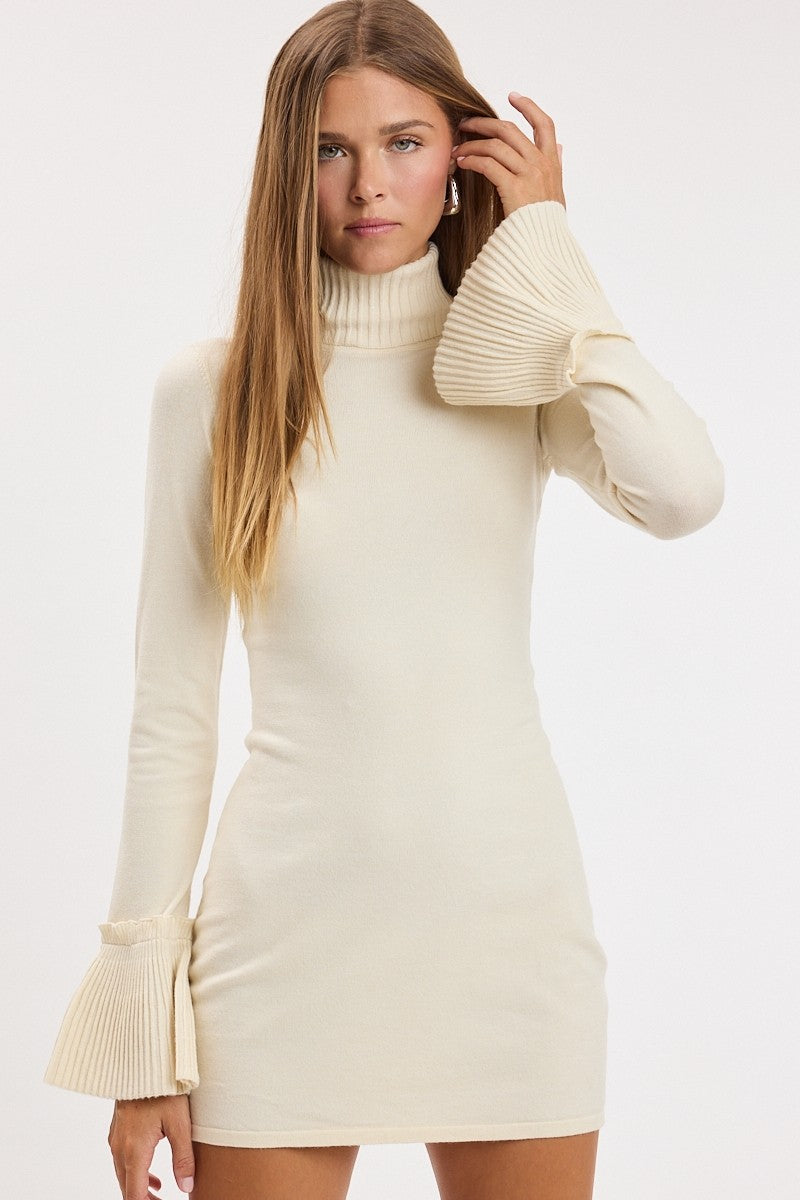 A woman modeling a cream-colored ribbed knit mini dress with a mock neck and dramatic pleated bell sleeves against a plain white studio background.