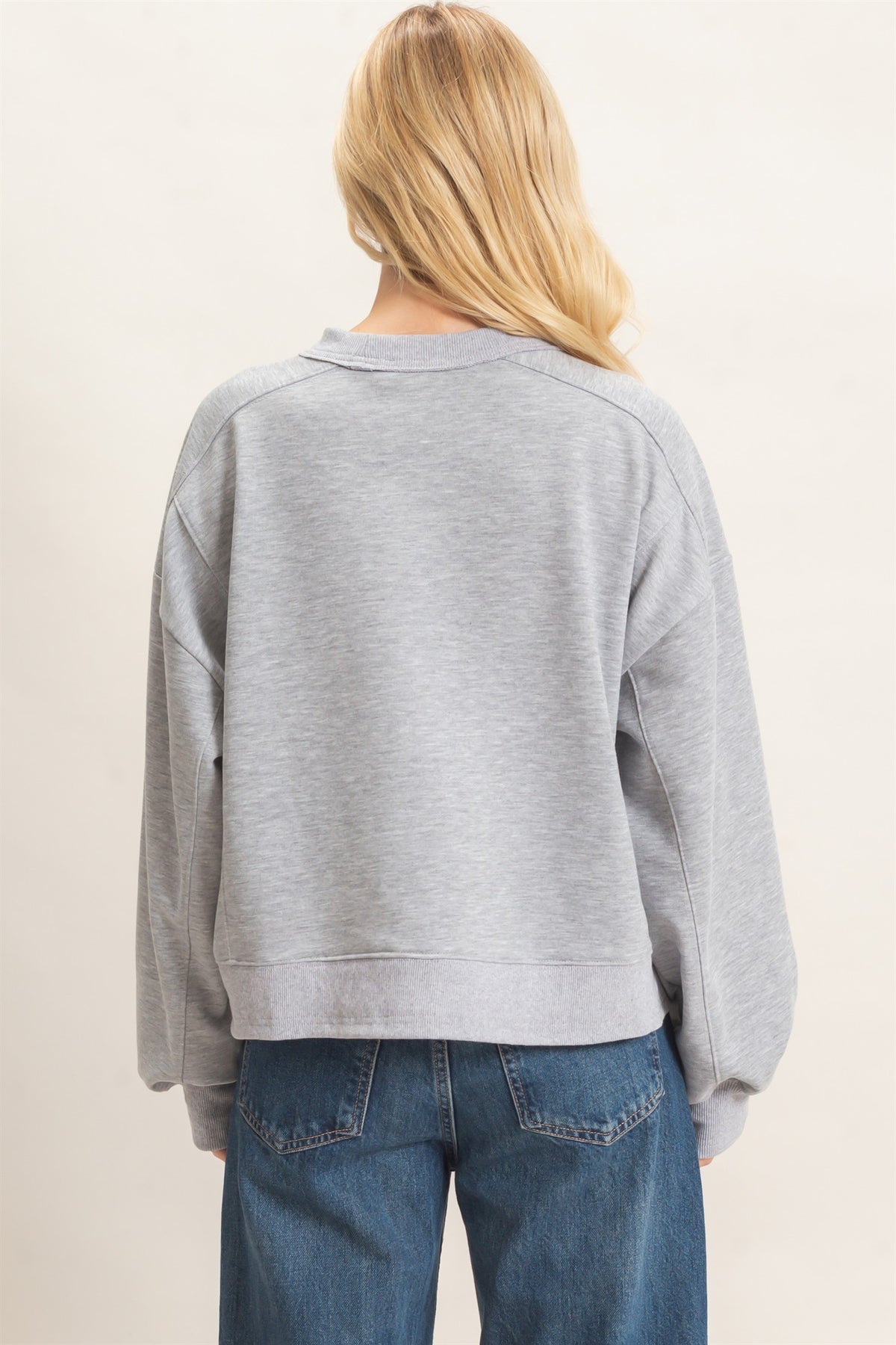 A woman viewed from behind wearing a light gray heather crewneck sweatshirt and blue jeans against a plain off-white background.