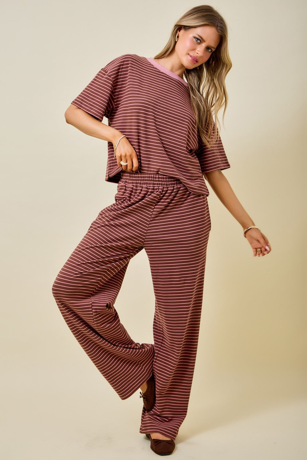A woman modeling a matching two-piece loungewear set consisting of a short-sleeve top and wide-leg pants, both featuring a dark red and brown horizontal stripe pattern, set against a solid beige background.