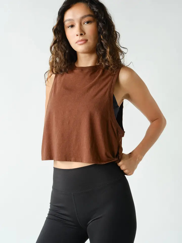 A woman modeling a sleeveless cropped tank top in a warm brown color, paired with black leggings, against a plain light gray studio background.